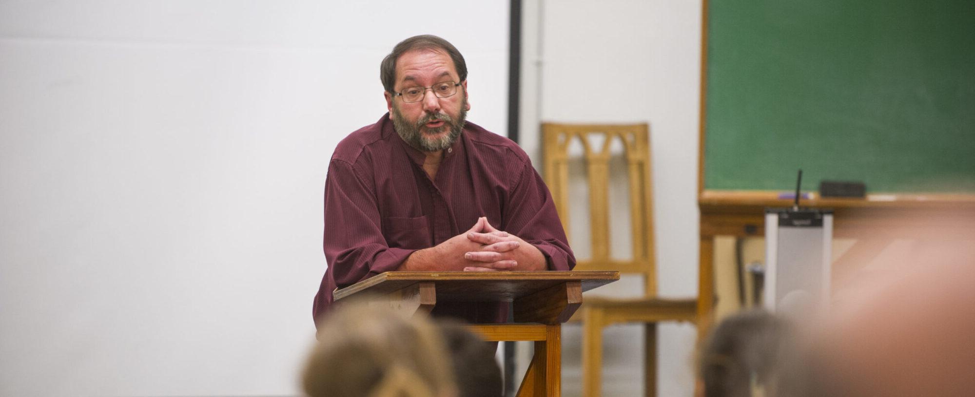 Man reading to a classroom from a podium.