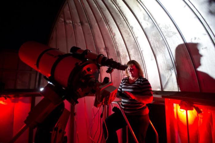 A woman observes the night sky using a telescope inside a dome observatory illuminated by red lighting.