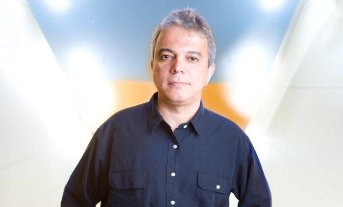 A man with gray hair wearing a navy-blue shirt poses confidently in a bright, softly lit space with gradient colors.