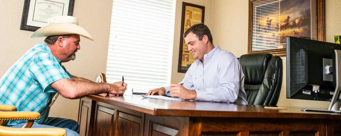 Two men signing documents in an office, one is sitting behind the desk with a computer on his side and the other is sitting in front wearing a hat. 