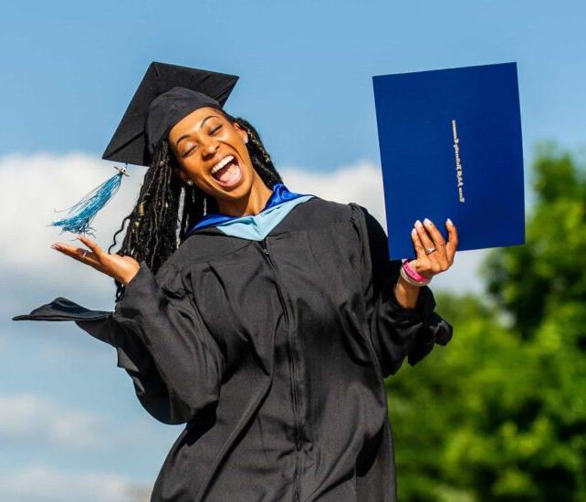 A joyful graduate in a black cap and gown smiles widely while holding up a blue diploma cover in one hand and gesturing with the other. The background features a clear sky and greenery.