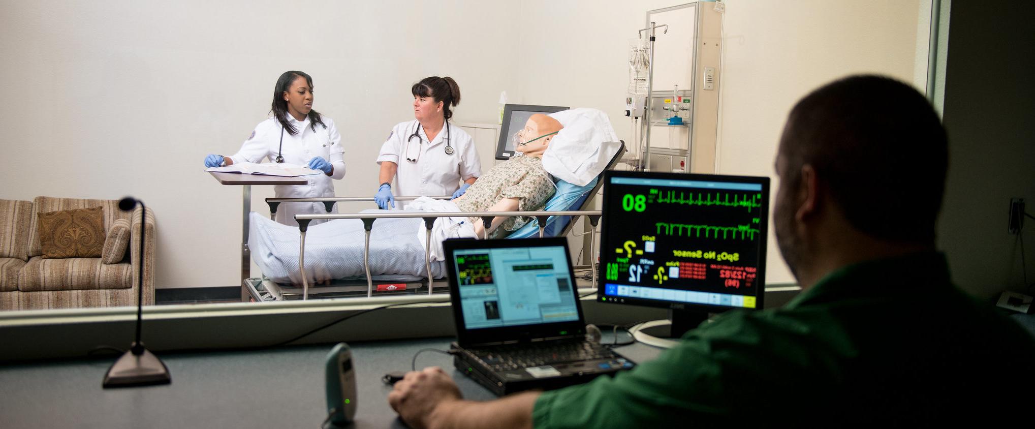 A man at computer behind computers with viewing window of nurses practicing on a mannequin.