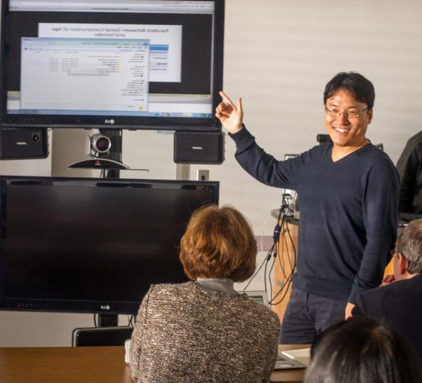 Male teacher point at TV screen and conferencing camera in front of class.