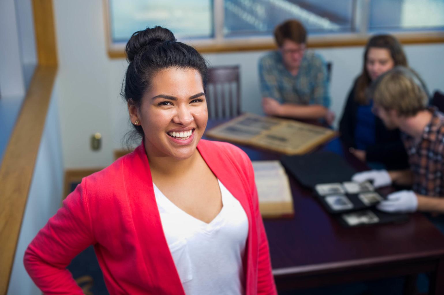 Young woman smiling with students looking at historic photographs in the background.