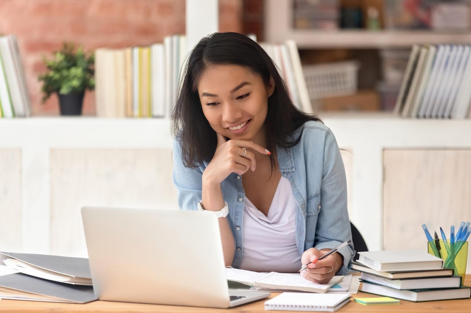 A applied criminology student sitting down at a desk, taking notes and looking at the screen of a laptop.