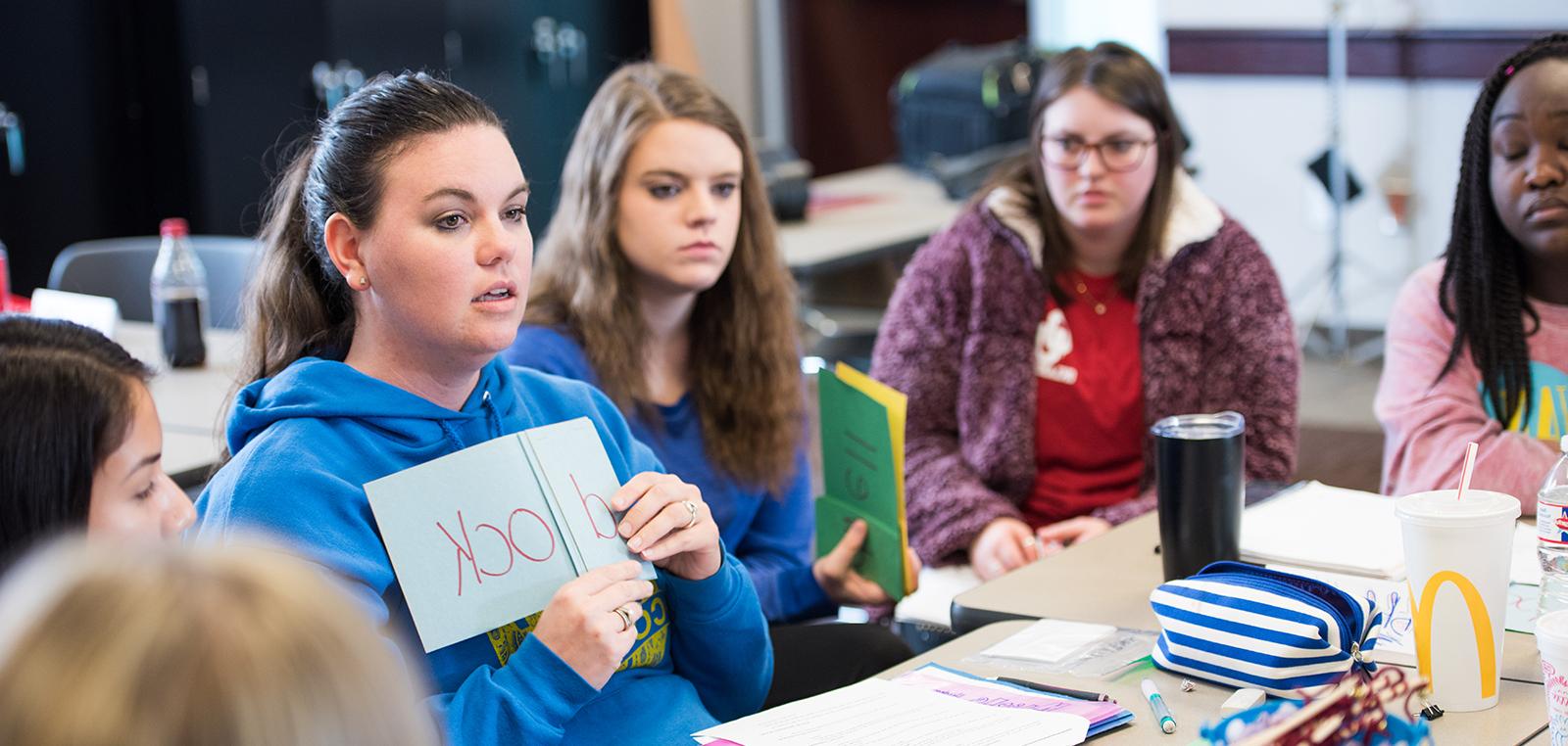 A student holding a blue piece of paper with a word wrote on it, in class.