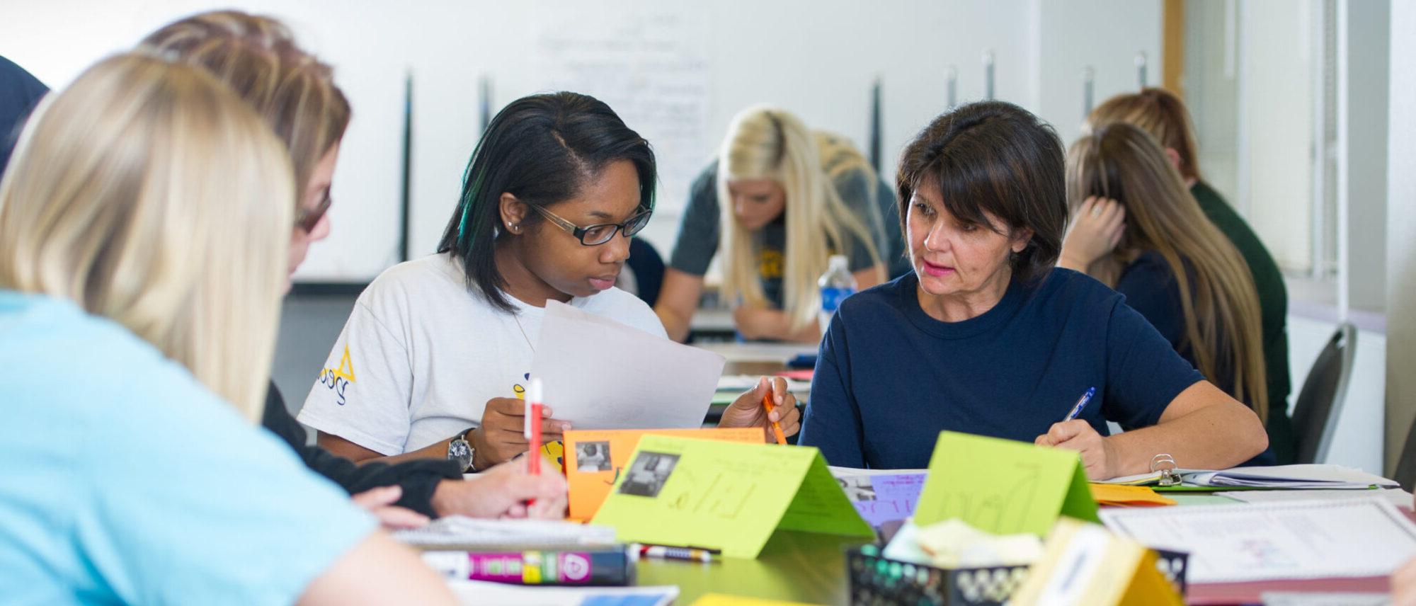 Two students working together while reading material on a desk.