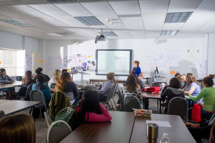 A classroom with a teacher standing at the front near a projected presentation. Students sit at tables, attentively facing the teacher, with posters on the walls.