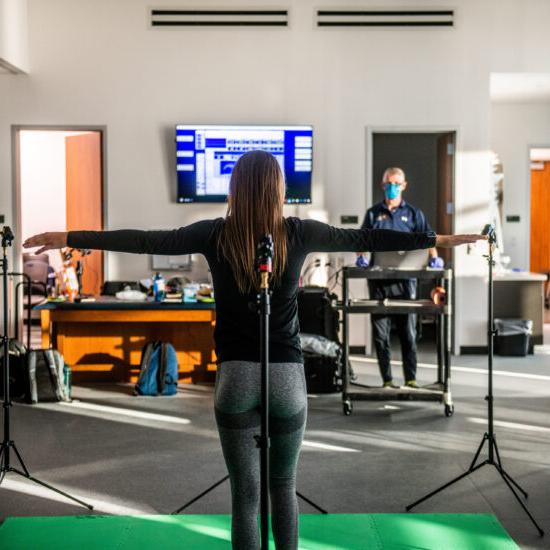 A person stands on a green mat with arms outstretched, facing a screen displaying data. A researcher in a mask observes and records information in a lab setting.