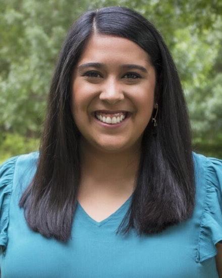 A smiling female with tanned skin and long dark brown hair. She is wearing a blue v neck blouse. In the background is trees.