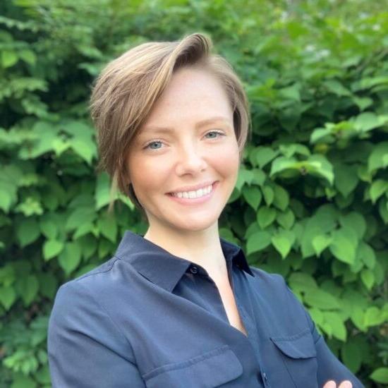 A smiling short hair dirty blonde female. She is wearing a black button up blouse. In the background is green trees.