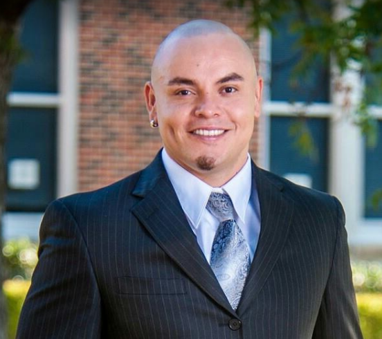 A bald young man smiling. He is wearing a black dress suit with a blue tie. In the background is a brick house with windows.