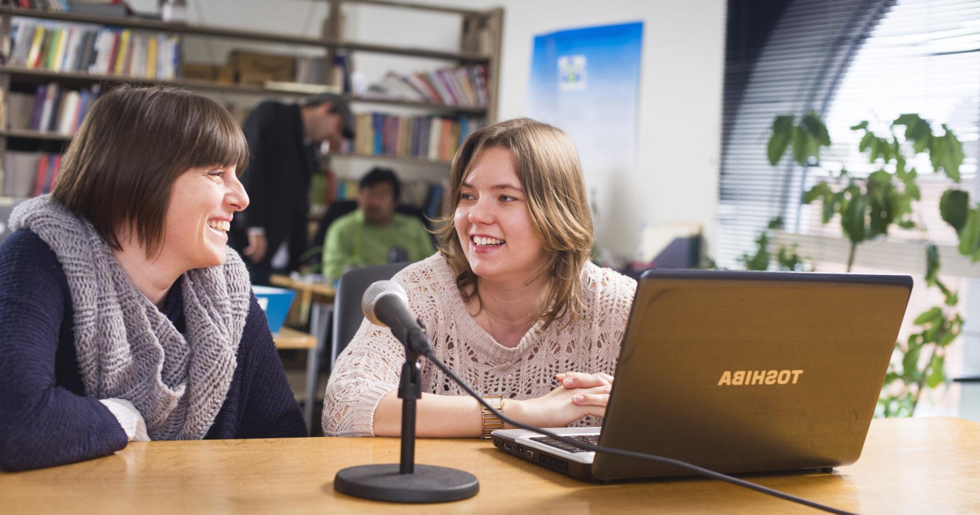 A professor and a student sitting down at a desk with a computer and a microphone in front of them.