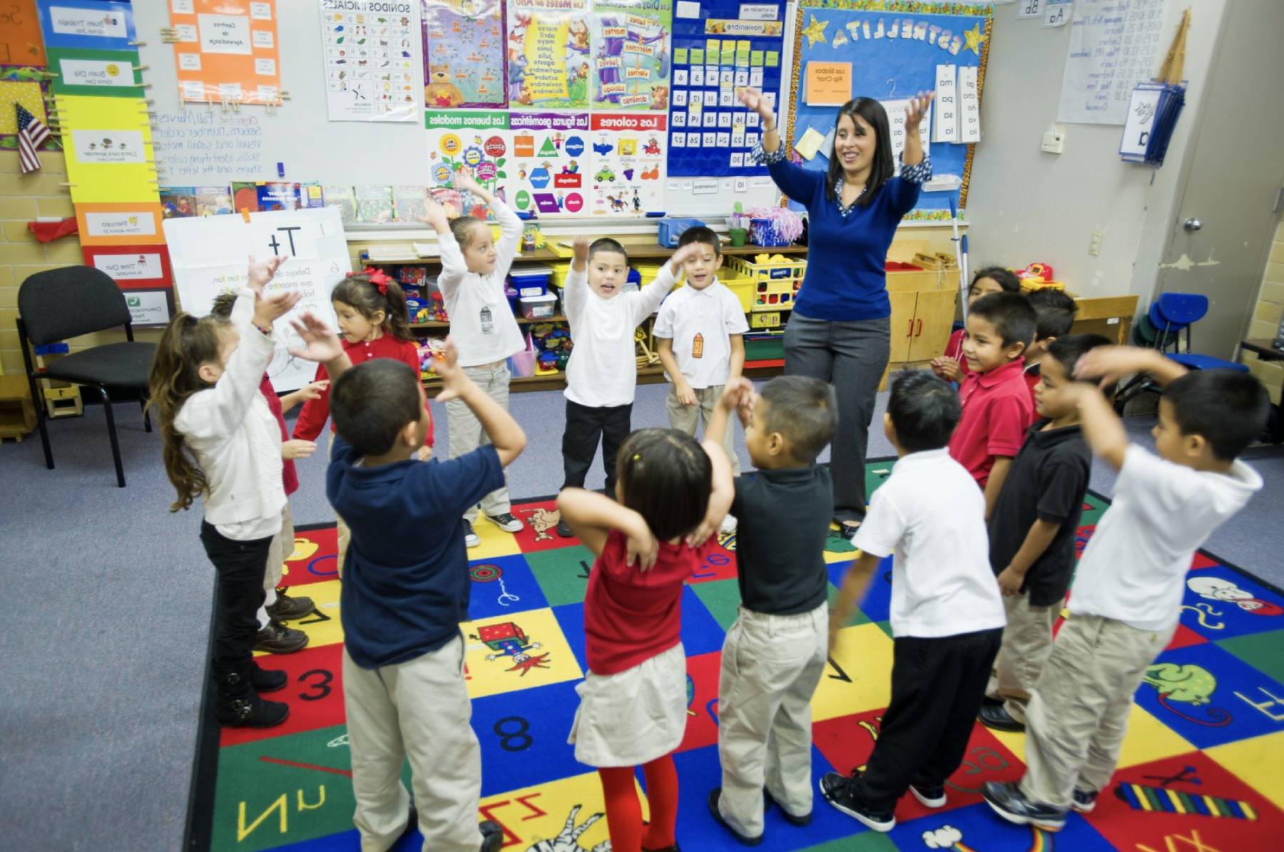 A teacher stands in front of a group of young students in a colorful classroom. The children are excitedly participating in an interactive lesson, raising their arms while standing on a vibrant alphabet rug.