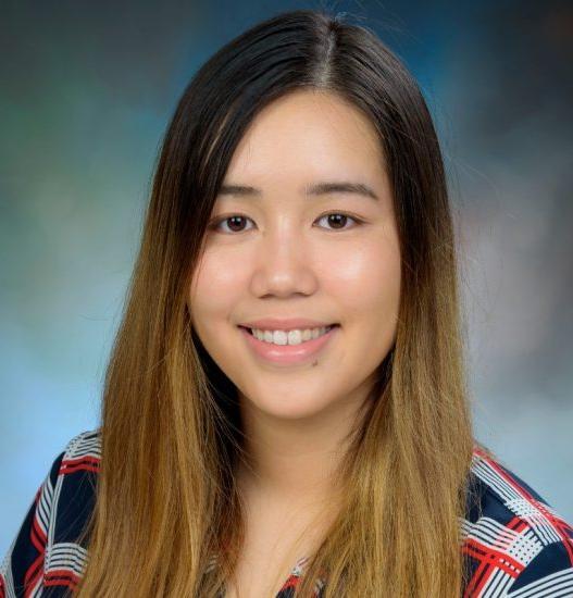 A young woman with long brown hair, wearing a plaid-patterned blouse, smiles for a headshot against a softly blurred blue and green background.