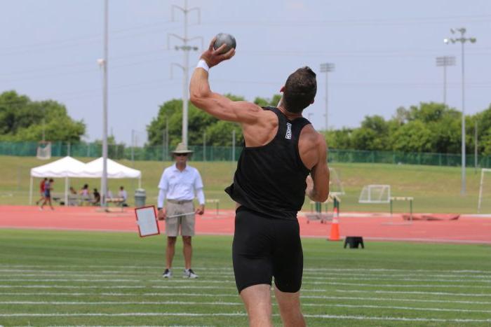 A male athlete competing in the shot put event launches a shot into the air.