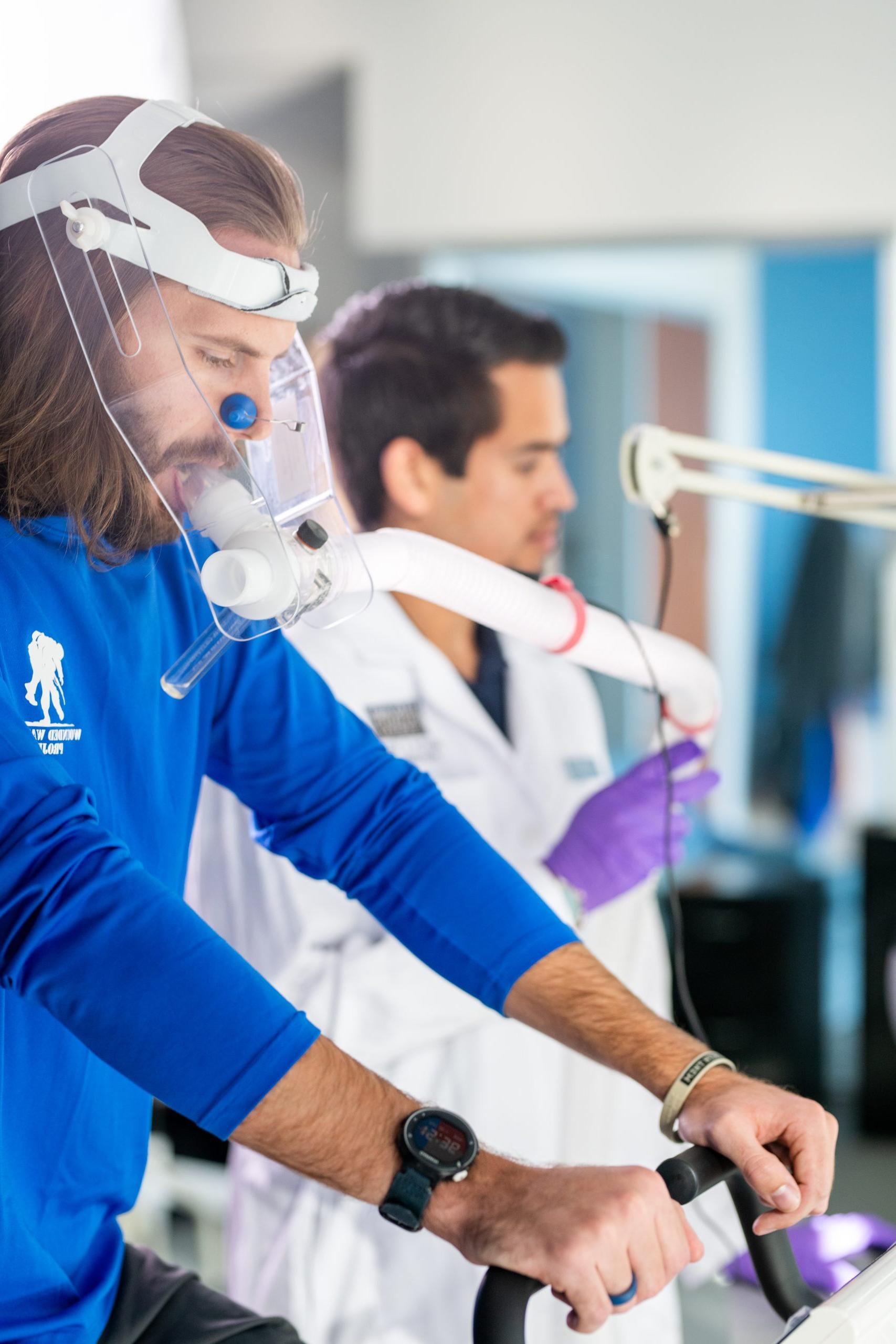 A man in a blue shirt cycles on an exercise bike wearing a breathing mask, monitored by a researcher in a lab coat. The setting is a scientific or medical research facility.