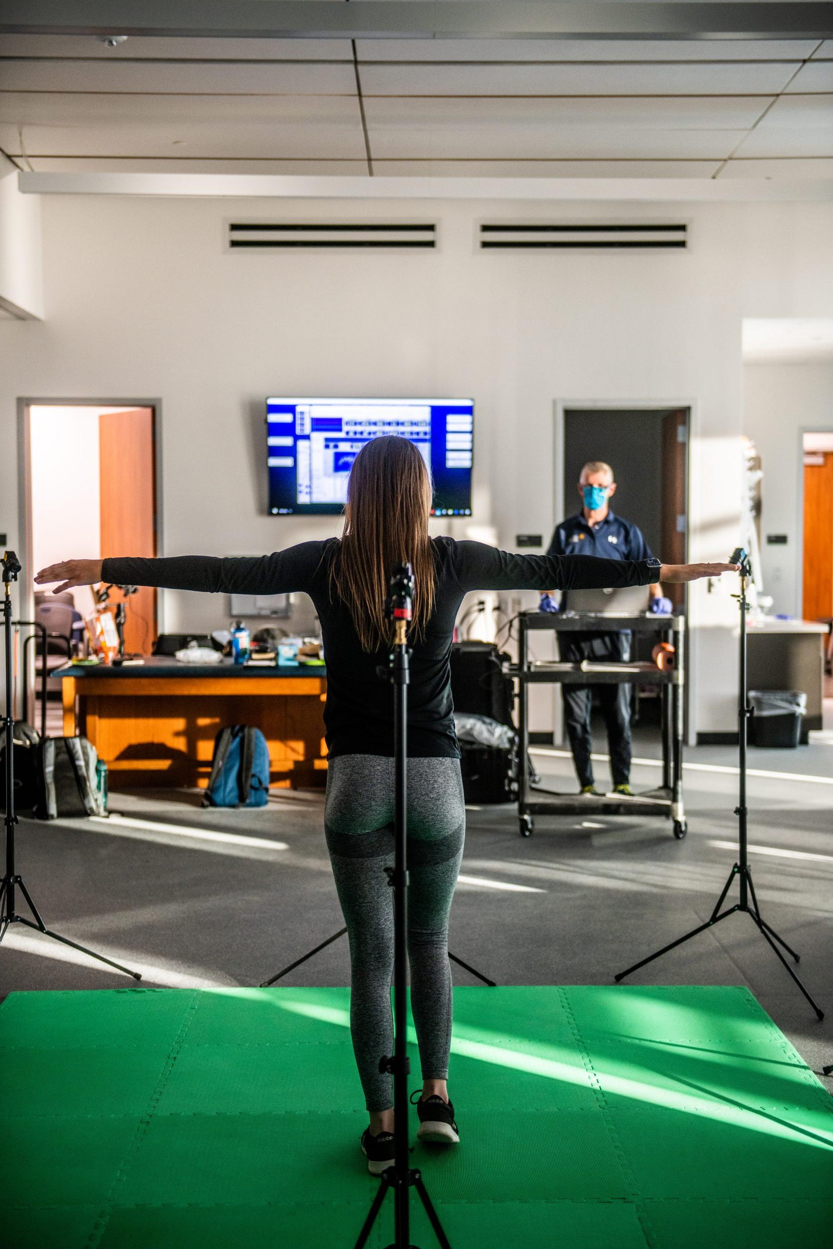 A woman stands facing away with arms raised as part of a physical assessment conducted by a faculty researcher.