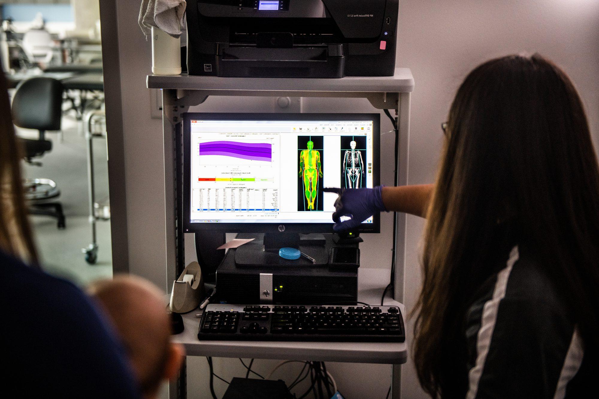 A researcher in gloves points at a computer screen displaying a full-body scan and data analysis, likely in a medical or scientific setting, with another person observing nearby.