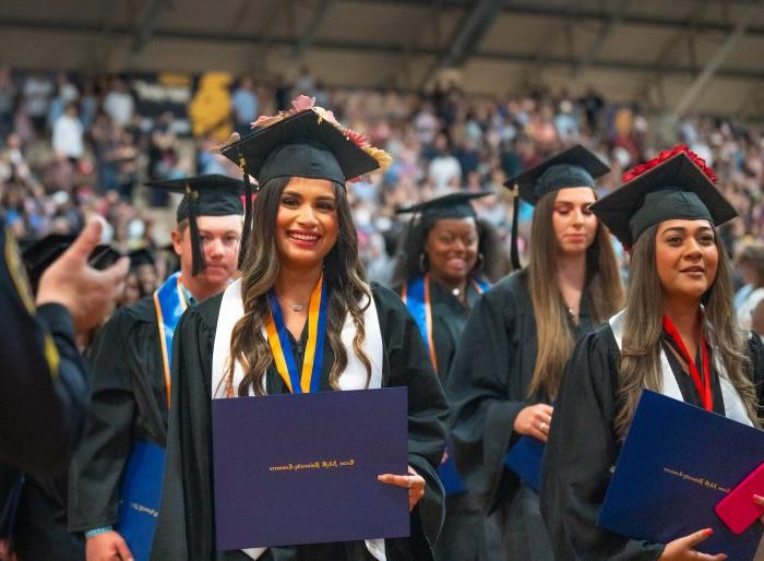 Graduates stand with their caps and gowns. Graduate in the foreground smiles with diploma in hand.