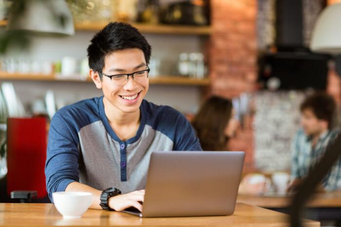 Happy cheerful young asian male in glasses smiling and using laptop in cafe