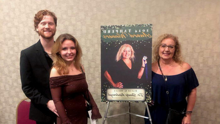 At left, Dr. Sandy Kimbrough stands next to a 2024 TAHPERD Award Winners sign showing a picture of her. On the right side of the image stands a female student with a male student behind her.