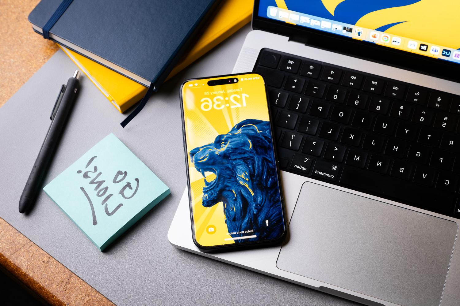 Close-up of a desk featuring a laptop, smartphone with a lion-themed wallpaper, and a note reading 'Go Lions!